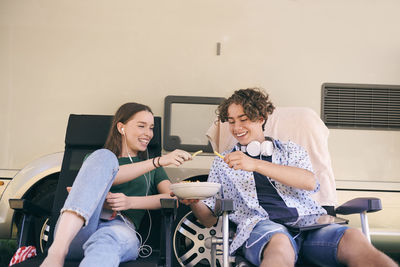 Cheerful siblings enjoying snacks while sitting on chairs against van at campsite