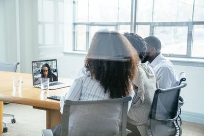 Business people video conferencing with businesswoman over laptop computer in board room seen through glass