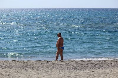 Side view of shirtless overweight man standing on shore at beach during sunny day