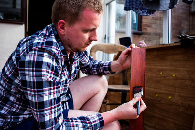 Side view of young man painting wooden plank while crouching at home