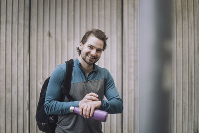 Portrait of happy young man with backpack and water bottle standing against wall