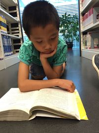 Boy reading book while kneeling by bookshelf