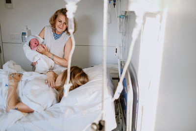 Happy mature female greeting newborn grandchild in hospital ward while young mother lying in bed and receiving medical treatment