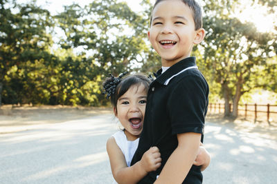 Portrait of happy sister with mouth open embracing brother while standing on road against trees in park