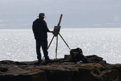 Rear view of mature man painting while standing on rock by sea against sky during sunny day