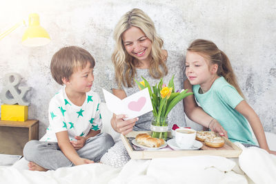 Smiling woman reading greeting card while children looking at her on bed