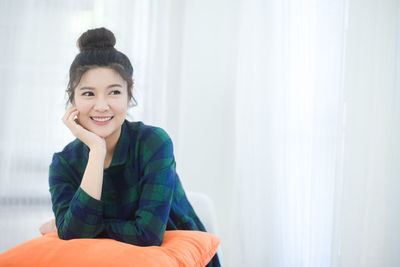 Smiling woman with orange pillow sitting against wall at home