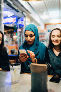 Young muslim woman using smart phone while sitting amidst female friends in cafe