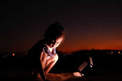 Rear view of boy sitting on land at beach against sky at night