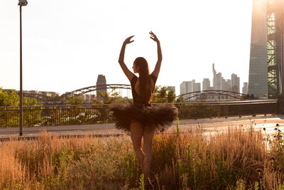 Rear view of female ballet dancer dancing on grassy field in city against clear sky