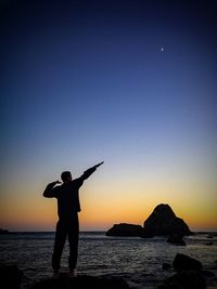 Rear view of man gesturing at sea shore against sky during sunset