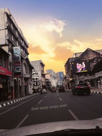Cars on road by buildings against sky during sunset