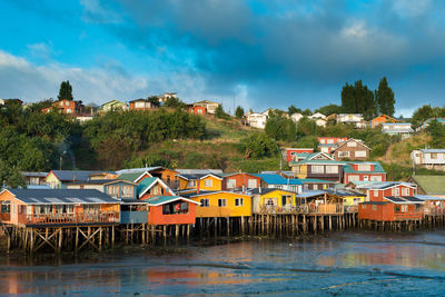Houses by river and buildings against blue sky