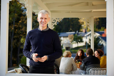 Portrait of confident smiling mature man standing on porch with friends in background