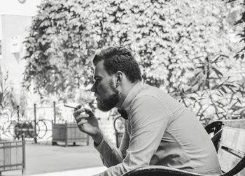 Side view of young man smoking cigarette while sitting on bench in city