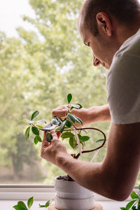 Young man cutting potted plant by window at home