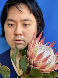 Close-up portrait of young asian man with pink king protea flower against blue background.