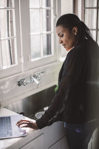 Side view of female professional using laptop while standing by sink at home office