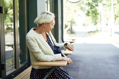 Side view of trendy female sitting at table in street cafe and checking time on wristwatch while waiting for appointment