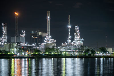 Illuminated factory by river against sky at night