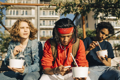 Young man using mobile phone while friends eating take out food in city during sunny day