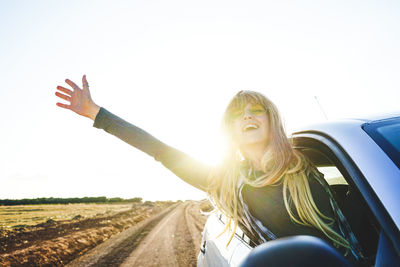 Young woman with arms outstretched leaning out from car window against clear sky