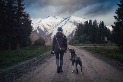 Rear view of woman with dog walking on dirt road leading towards mountains