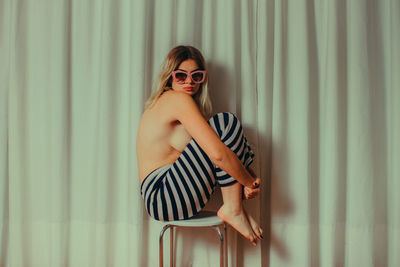 Side view of shirtless young woman wearing sunglasses while sitting on chair by curtain