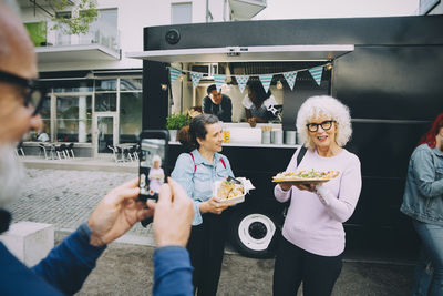 Senior man taking photograph of partner with indian food plate against commercial land vehicle