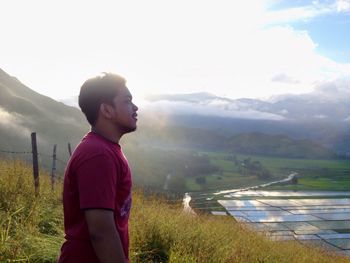 Side view of man standing on hill by farm against sky