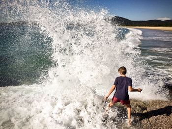 Rear view of boy standing by splashing water at beach 