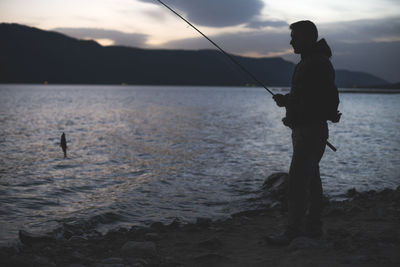 Silhouette man fishing in lake against sky at dusk