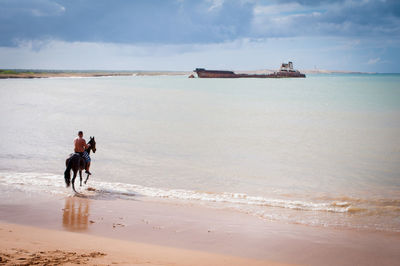 Shirtless man riding horse at beach against cloudy sky during sunny day