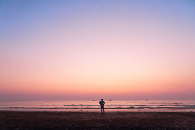 Rear view of man standing at beach against sky during sunset