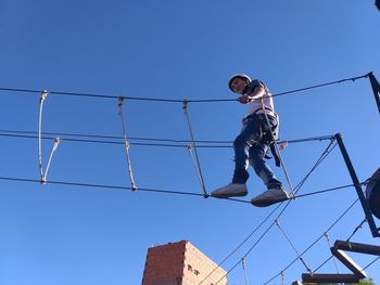 Low angle view of teenager walking on rope bridge against blue sky
