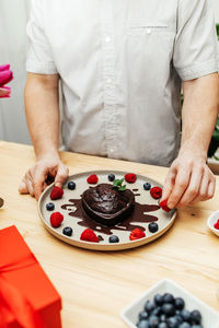 A man decorates a chocolate cake heart with fresh berries and chocolate icing. present, surprise 