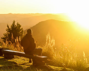 Rear view of woman sitting on mountain against sky during sunset