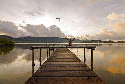 Rear view of man with arms raised sitting on pier over lake against during sunset
