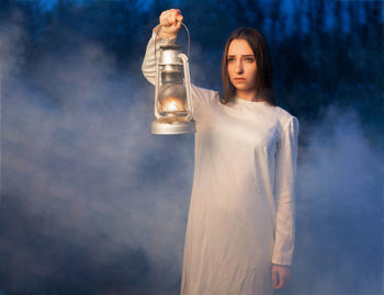 Young woman with illuminated oil lamp standing in forest at dusk