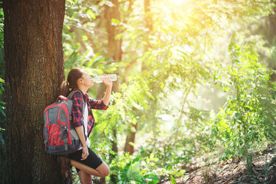 Female hiker drinking water while leaning on tree trunk in forest