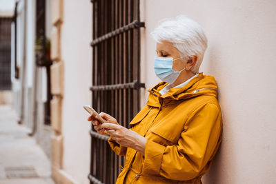 Side view of woman using mobile phone while standing against wall