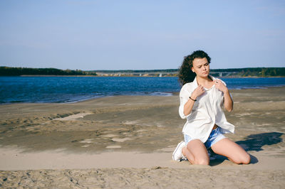 Beautiful young woman kneeling on sand against sea at beach