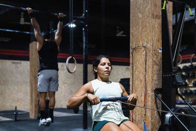 Strong hispanic sportswoman in activewear training on row machine during workout in light gym near sportsman handing on crossbar