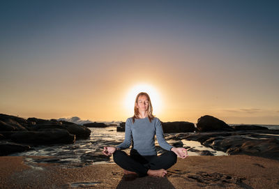 Woman meditating yoga on shore at beach against sky during sunrise