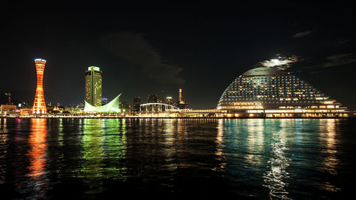 Illuminated modern buildings by river against sky at night