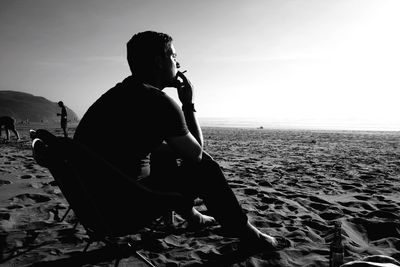 Full length of man smoking while sitting on chair at sandy beach against sky