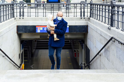 Man with face mask carrying baby while walking on staircase in city