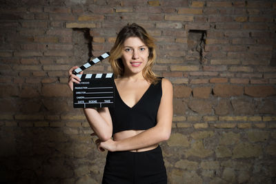Portrait of young woman holding film slate against brick wall