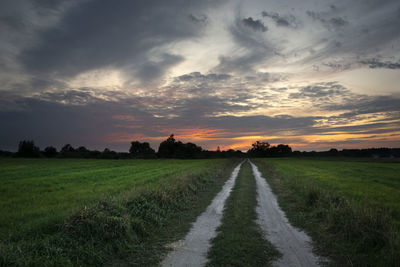 Dirt road and around green fields, clouds after sunset on the sky