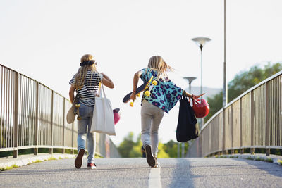 Full length of friends holding skateboards while walking on bridge against clear sky